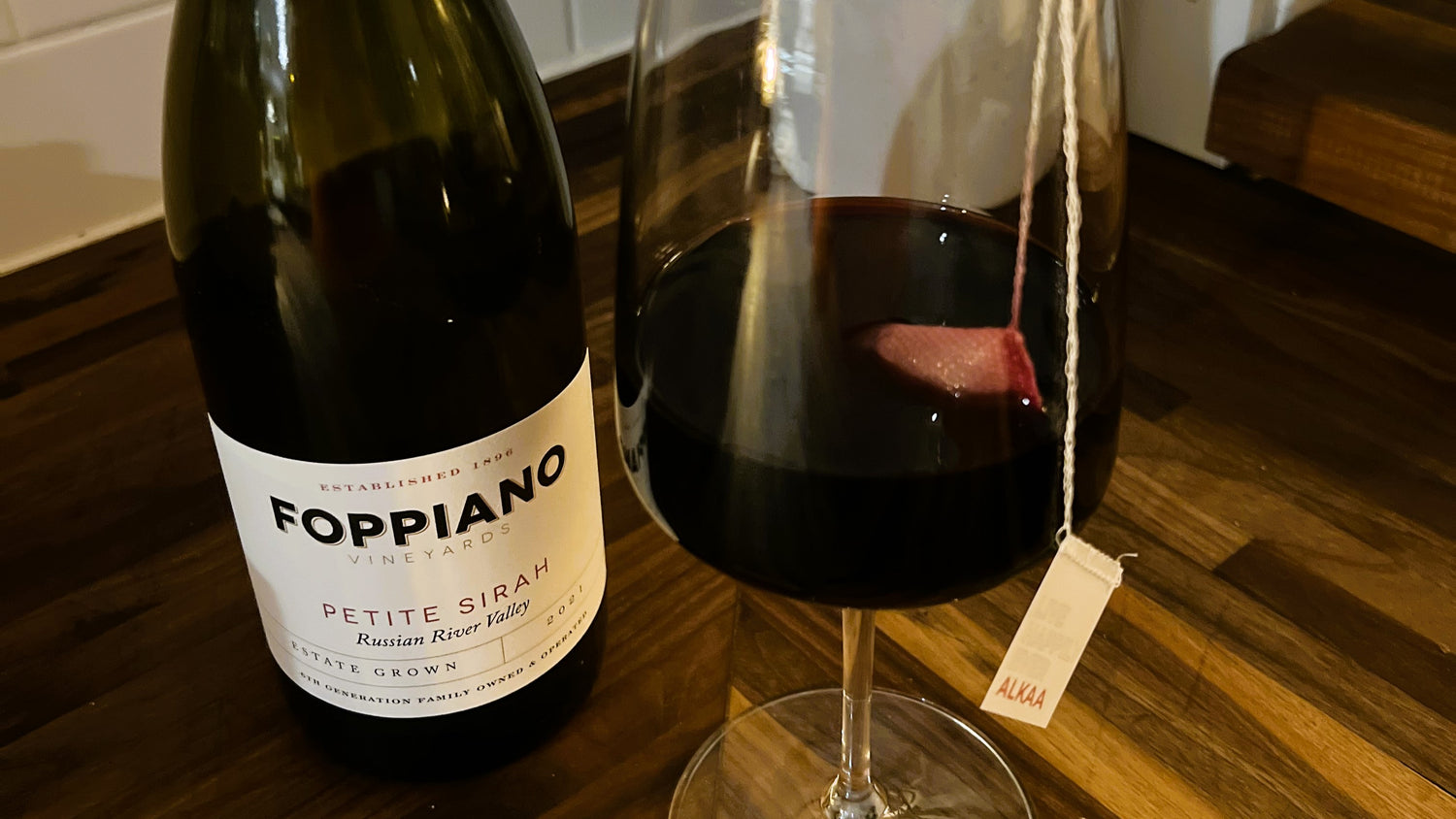 A Tagged ALKAA Sachet treating a glass of Foppiano Petite Sirah Wine on a butcher block counter.